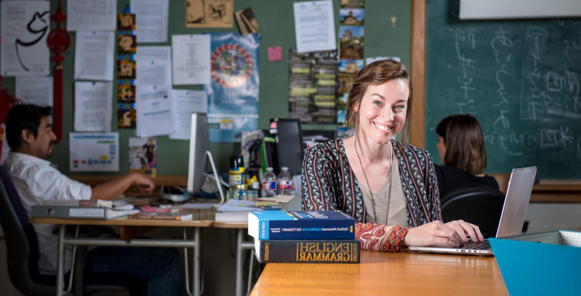 Young woman smiling at desk with laptop and English grammar books.
